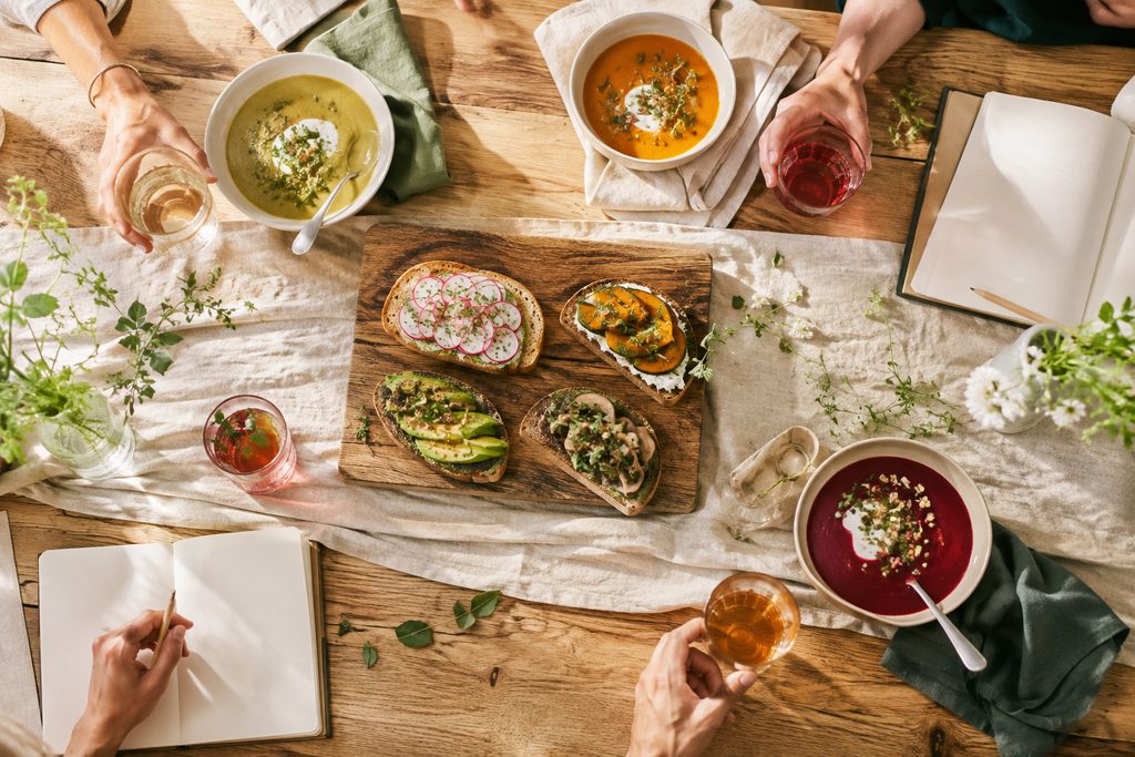 Grote gedeelde tafel, licht, waar men eet en praat rond dranken en kleine gerechten.